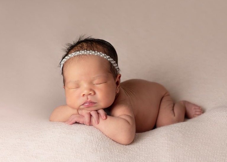 newborn baby girl with headband on blush pink blanket with chin on hands