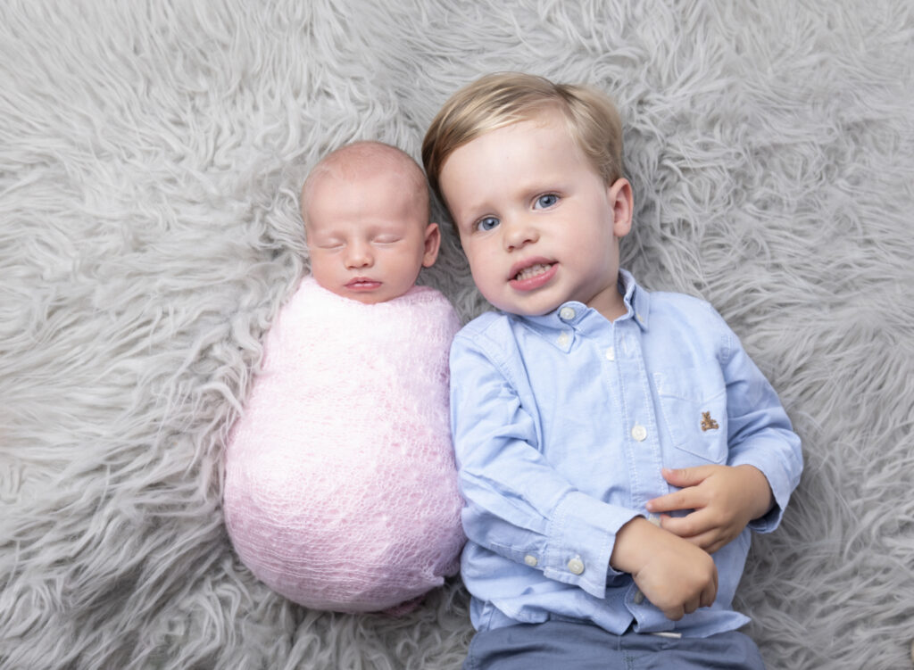 toddler boy with blond hair wearing a blue shirt laying next do his newborn baby sister who is wrapped in a pink lace wrap