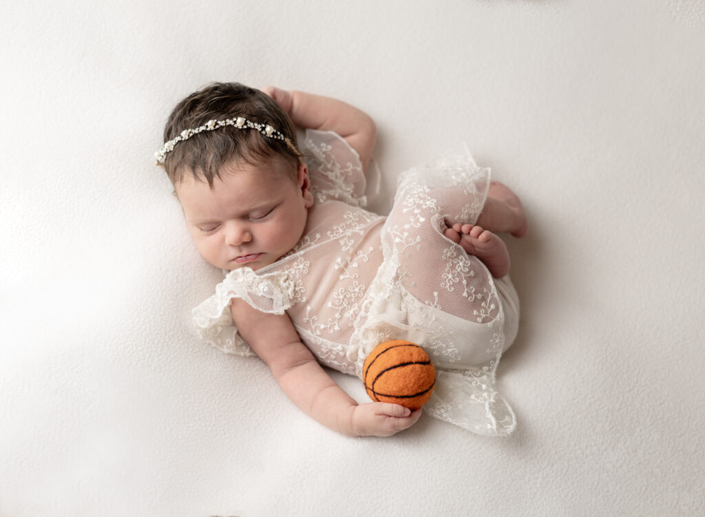 newborn girl wearing lacey dress and diamond headband posing with a miniature basketball