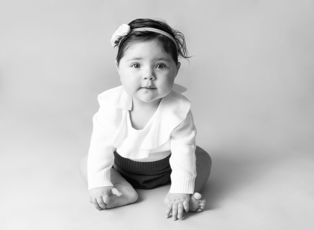 black and white image of baby girl sitting up in knit outfit with bow in her hair