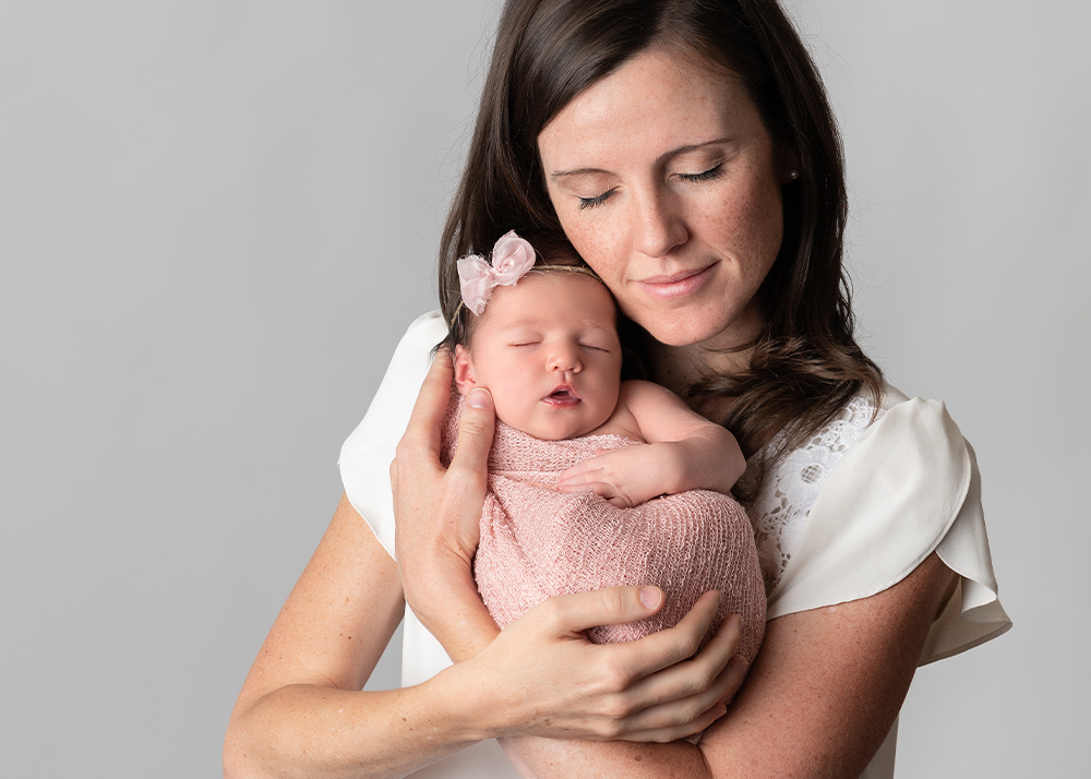 mom with her eyes closed holding her newborn daughter who is wearing a pink wrap and pink bow with their faces next to each other