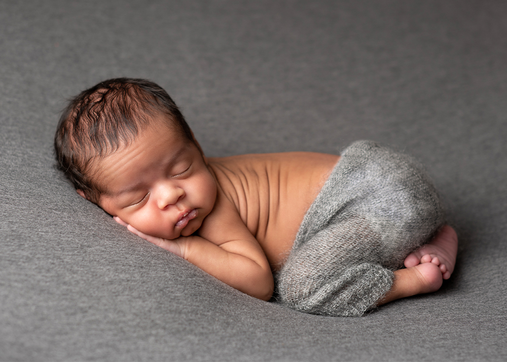 Newborn boy laying on his belly with his arms and legs tucked up under him and his head turned to the side resting on his hands on a medium gray backdrop