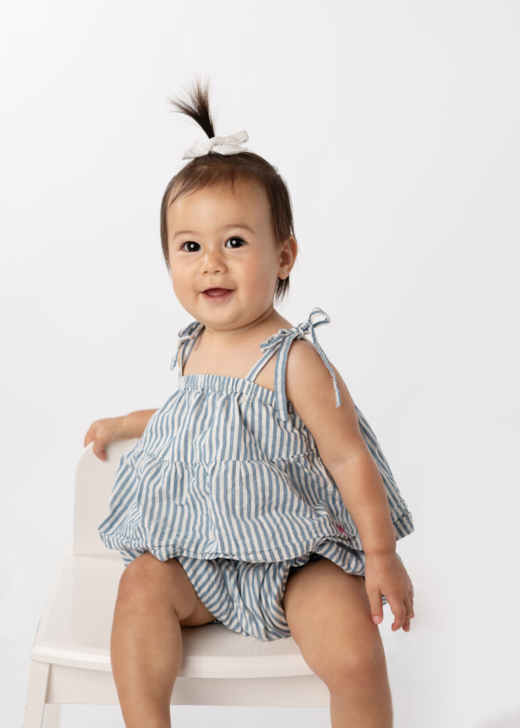 toddler girl with a tiny ponytail on the top of her head sitting on a small chair smiling for her school picture