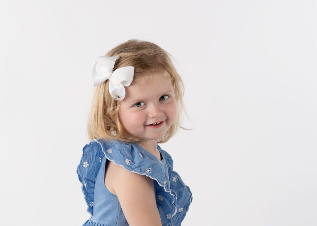 young girl with a denim dress and white bow smiling while looking back over her shoulder