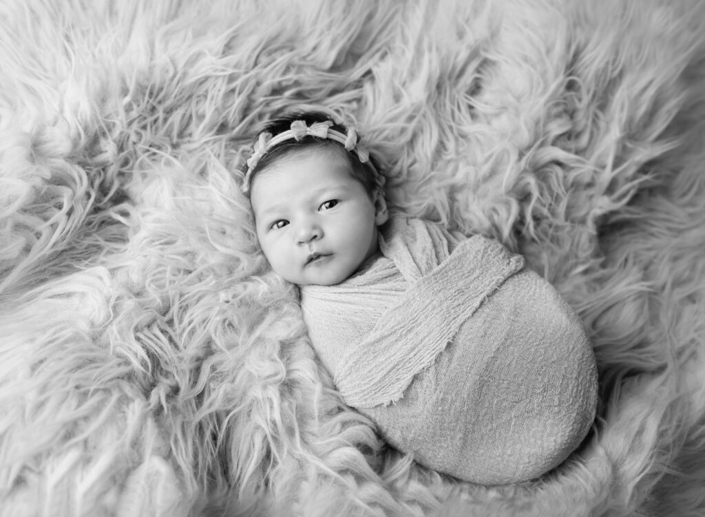 black and white image of newborn girl wrapped in swaddle and wearing a dainty headband on fur blanket