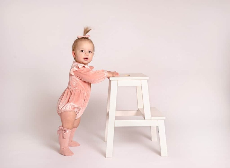 Annie standing with a stool for her 1st birthday milestone session in a pink velvet romper with knee high socks.