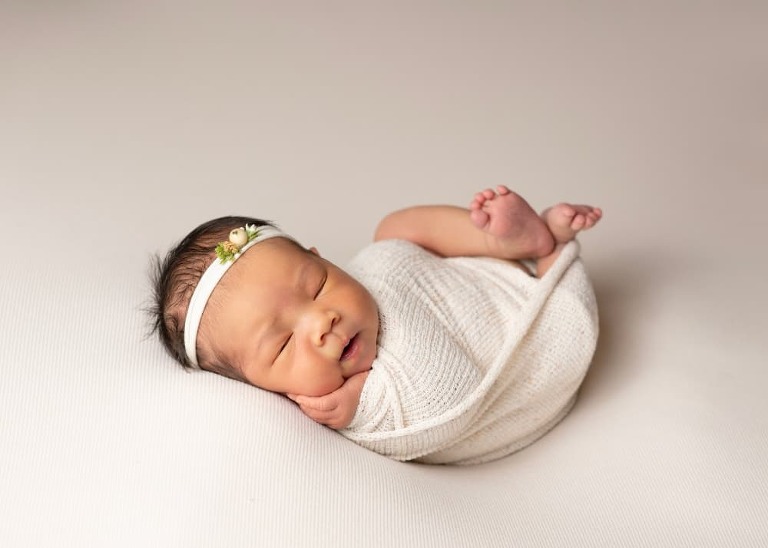 newborn girl in a cream wrap with a headband cupping her cheek with her hand